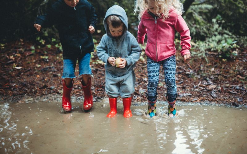 Drei Kinder mit Gummistiefeln spielen in einer Pfütze – zeigt, warum Wechselkleidung im Waldkindergarten wichtig ist.